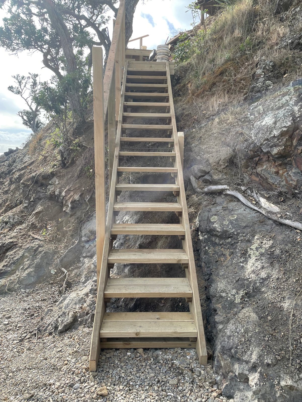 Finished timber staircase climbing a rocky hillside on Waiheke Island, set between stone walls and coastal vegetation