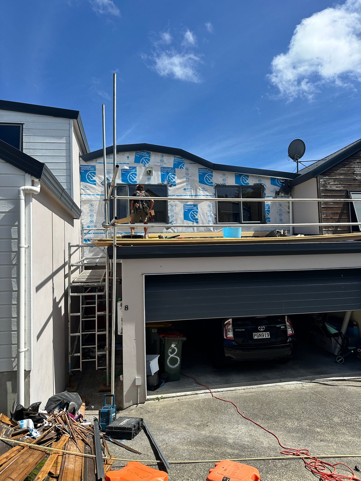 Two-story house under renovation with blue weatherproofing material covering upper level, open garage below, and construction materials in foreground.