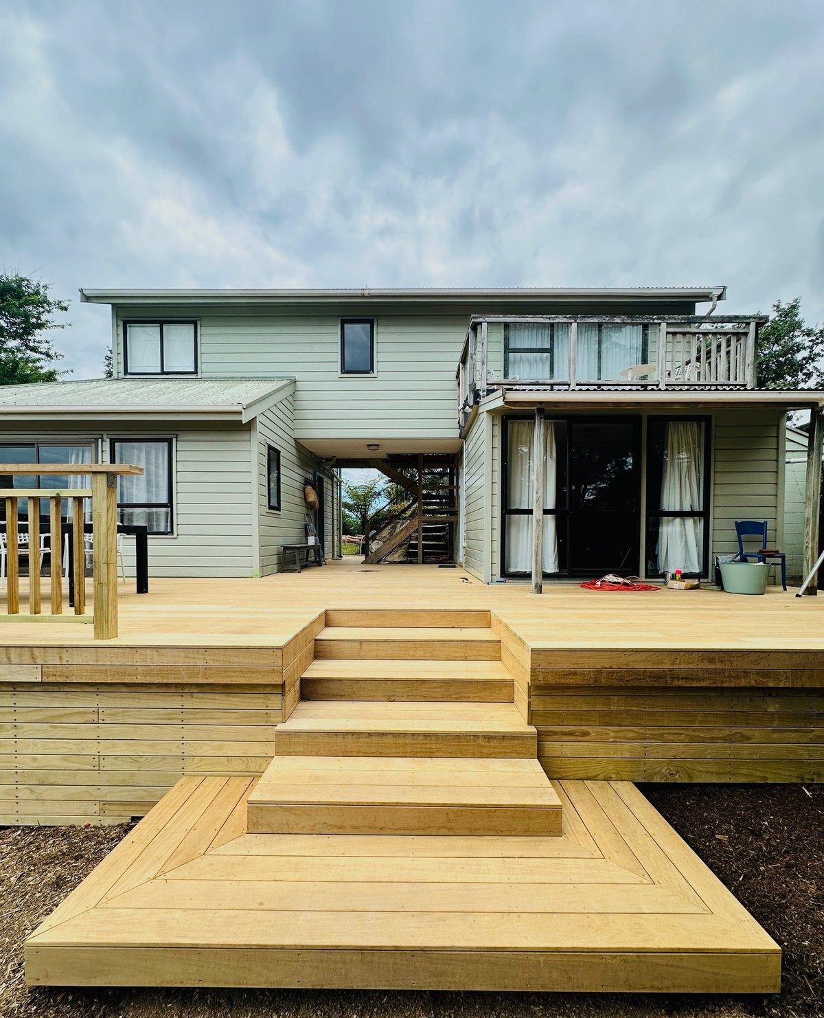 Raised wooden deck with stairs leading to a light blue single-story house with covered porch and deck under overcast sky
