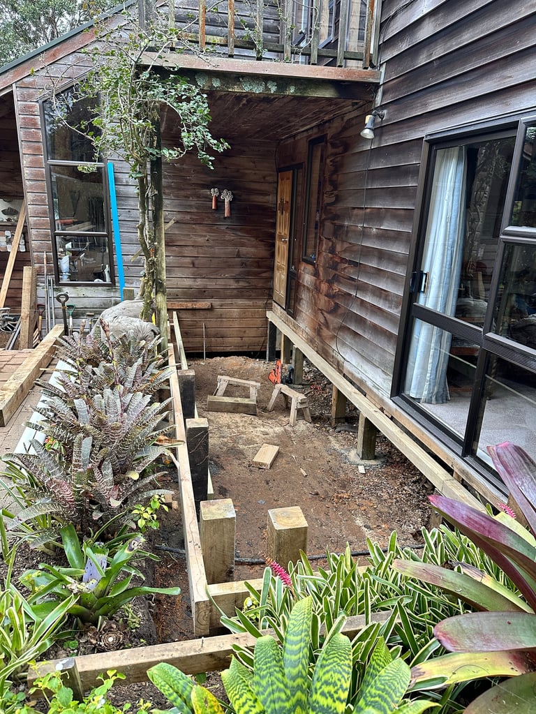 Narrow outdoor garden space between wooden houses with raised beds, stepping stones, and potted plants