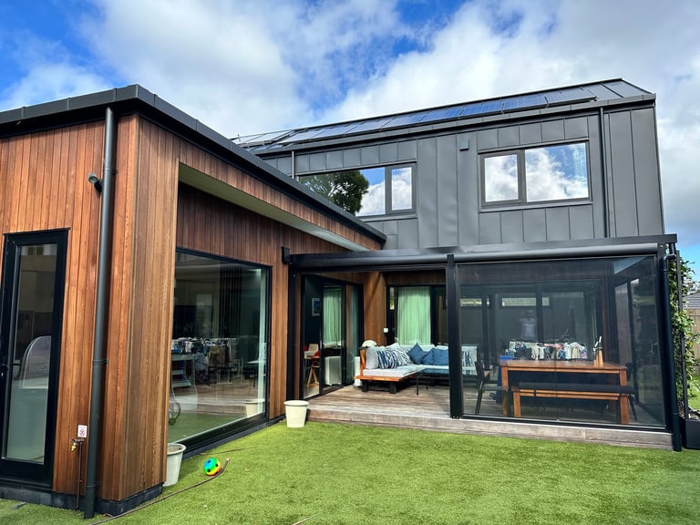 Modern two-storey home with warm vertical timber and dark metal exterior, large windows, open terrace and green lawn under a blue sky