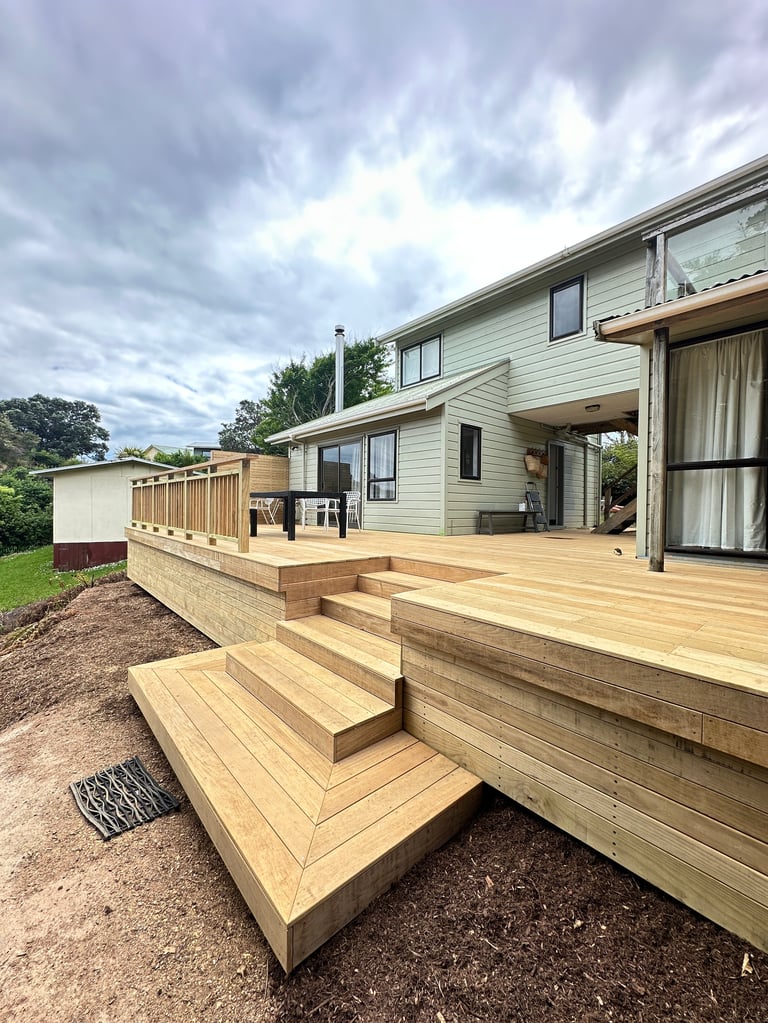 Modern beige house with wraparound wooden deck and stairs, surrounded by green lawn and trees under cloudy sky