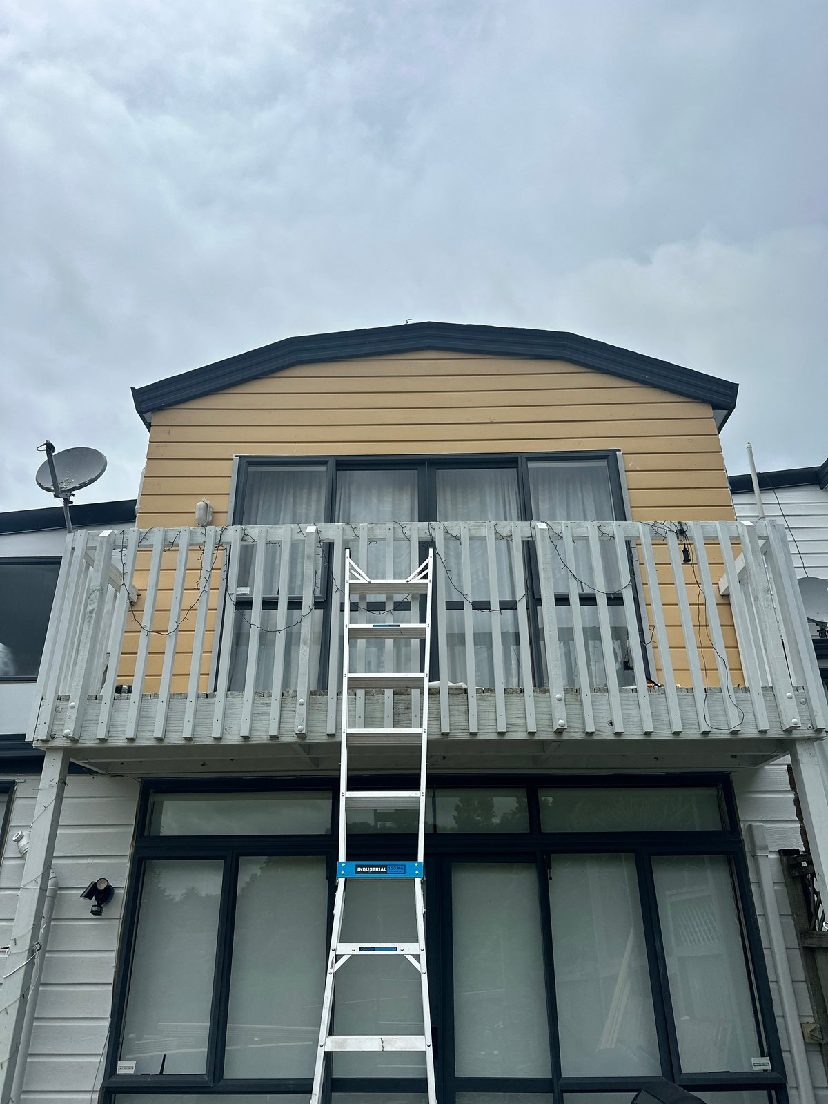 Modern two-story house with white railing deck and extended ladder leaning against the yellow upper level