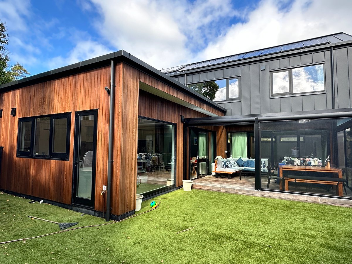 Modern two-story home with vertical timber and dark metal cladding, full-height glass doors, deck and green lawn under a blue sky
