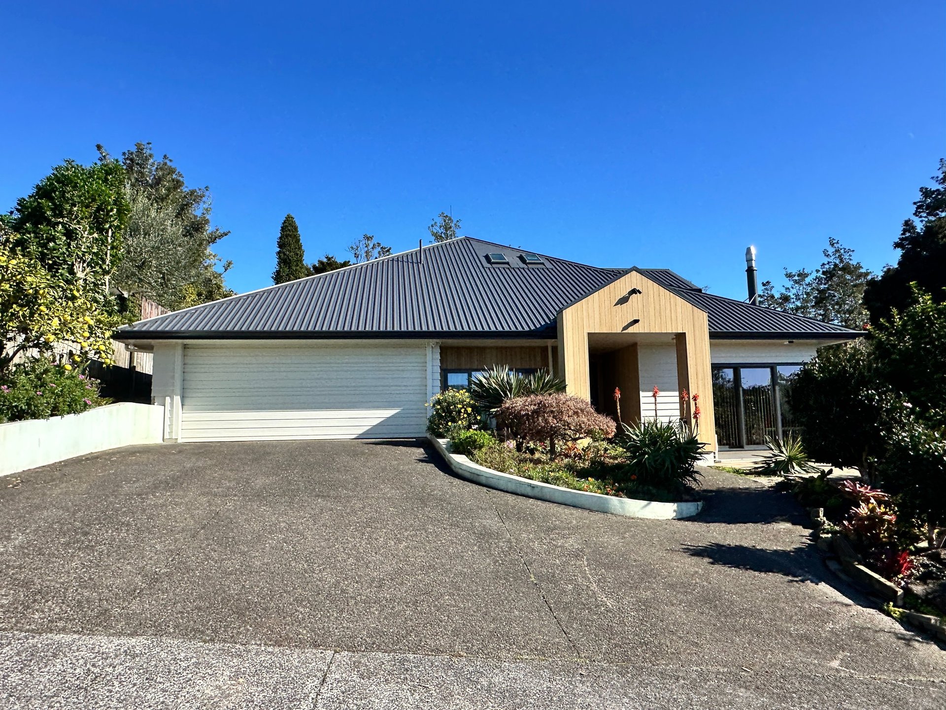 Modern single-story house with cream facade, dark metal roof, and white garage door set on a paved driveway with landscaped gardens and trees