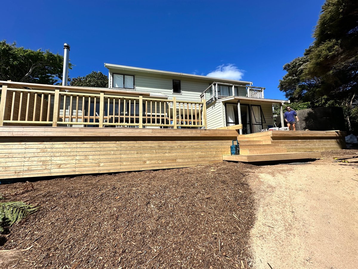 Modern single-story beach house with cream-colored railings and wooden deck under blue sky, surrounded by landscaping and sandy ground