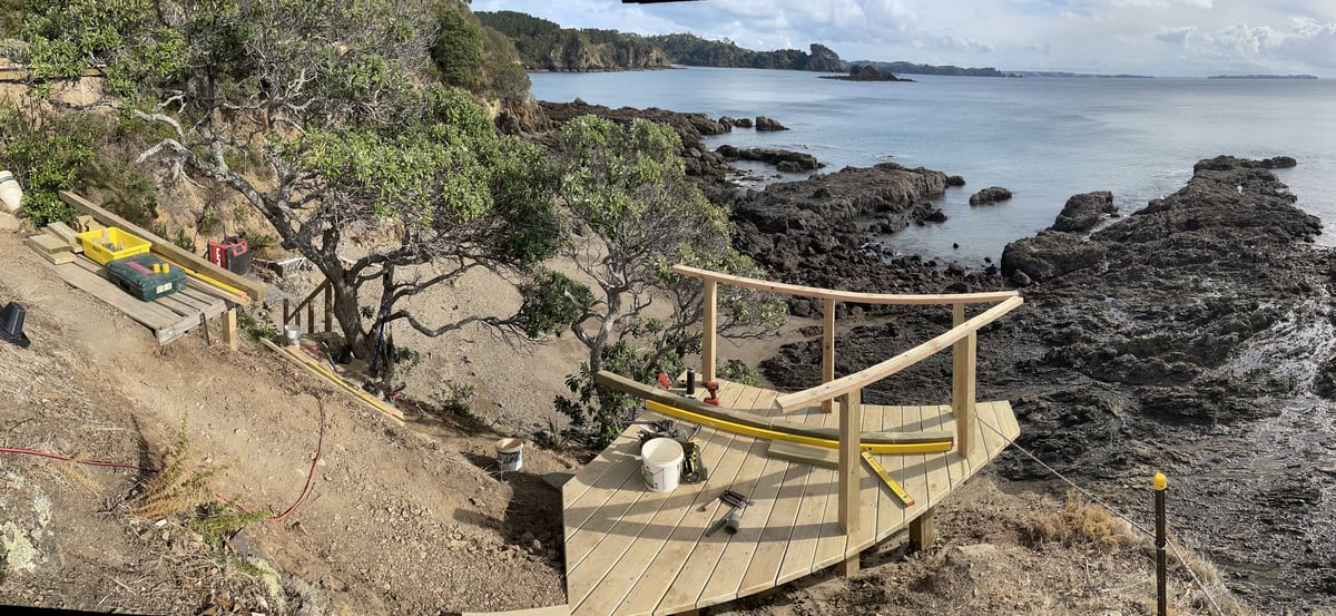 A timber corner stair flight built into a rocky cove, with a curved handrail set on intermediate posts and tools still on the deck