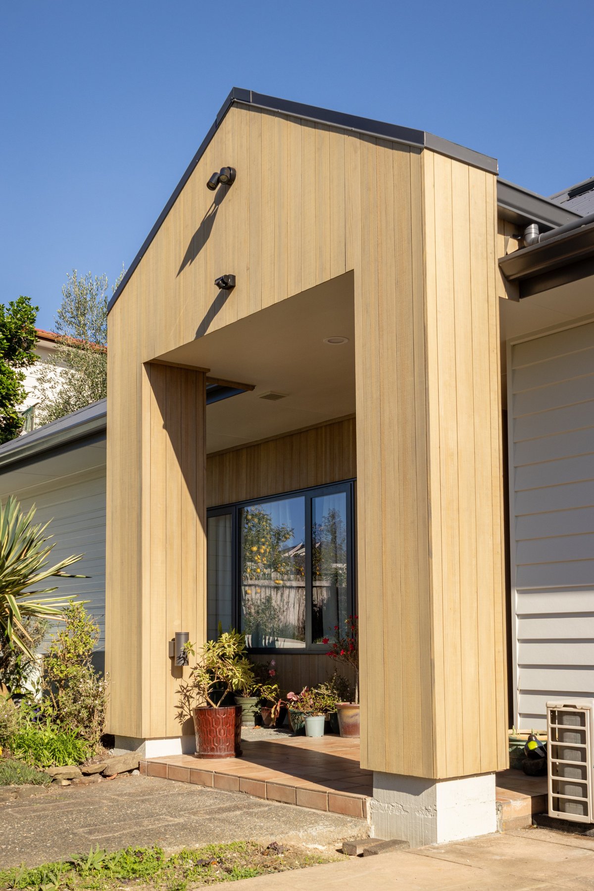 Modern wooden exterior addition with large window, metal hinges, and potted plants on windowsill