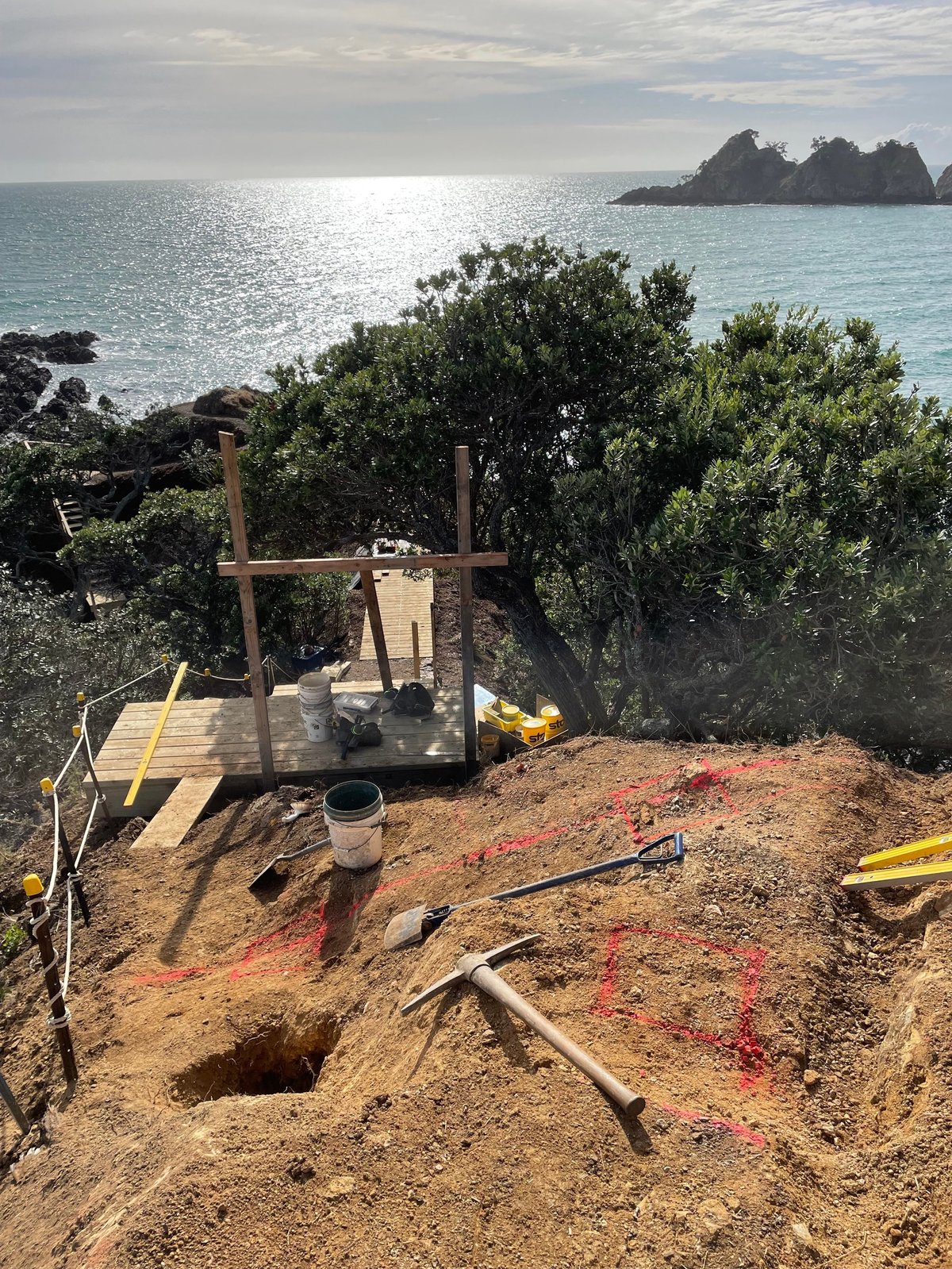 Coastal construction site with wooden frame structure, excavation hole, and tools on sandy beach overlooking turquoise ocean and rocky island