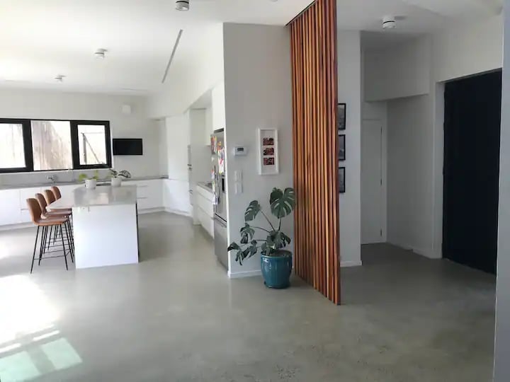 Modern open-plan interior with white kitchen island, vertical timber slat divider screen, potted plant and polished concrete flooring