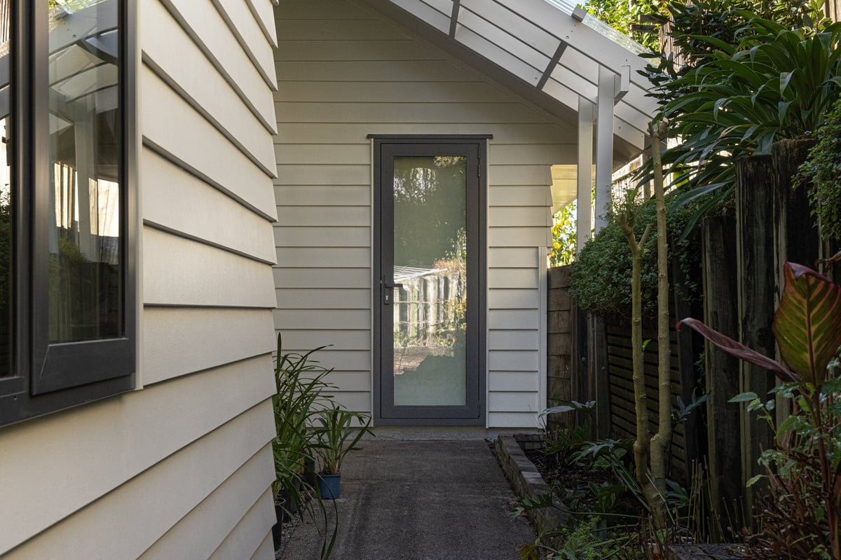 Modern house exterior with gray siding, dark frame door, and narrow side pathway lined with plants and wooden fence