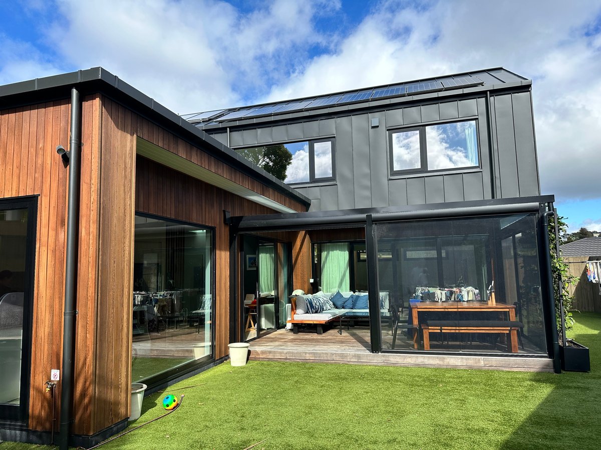 Modern black and timber home with full-height glass doors, covered deck, outdoor seating and manicured lawn under a blue sky