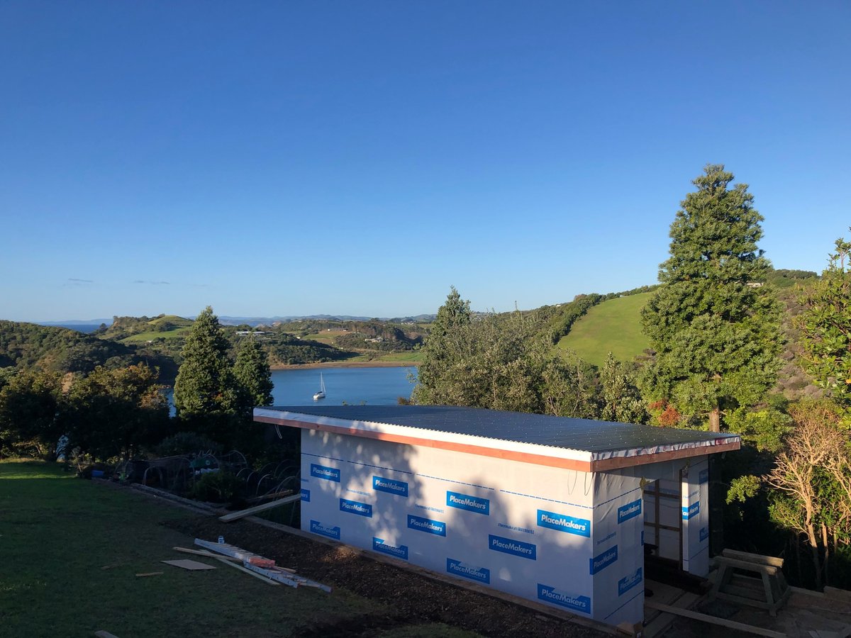 Modern blue and white cabin with sloped roof overlooking a lake, surrounded by green hills under a clear blue sky