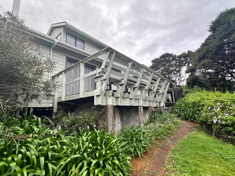 Elevated weathered house on wooden stilts surrounded by lush green gardens and trees under cloudy sky