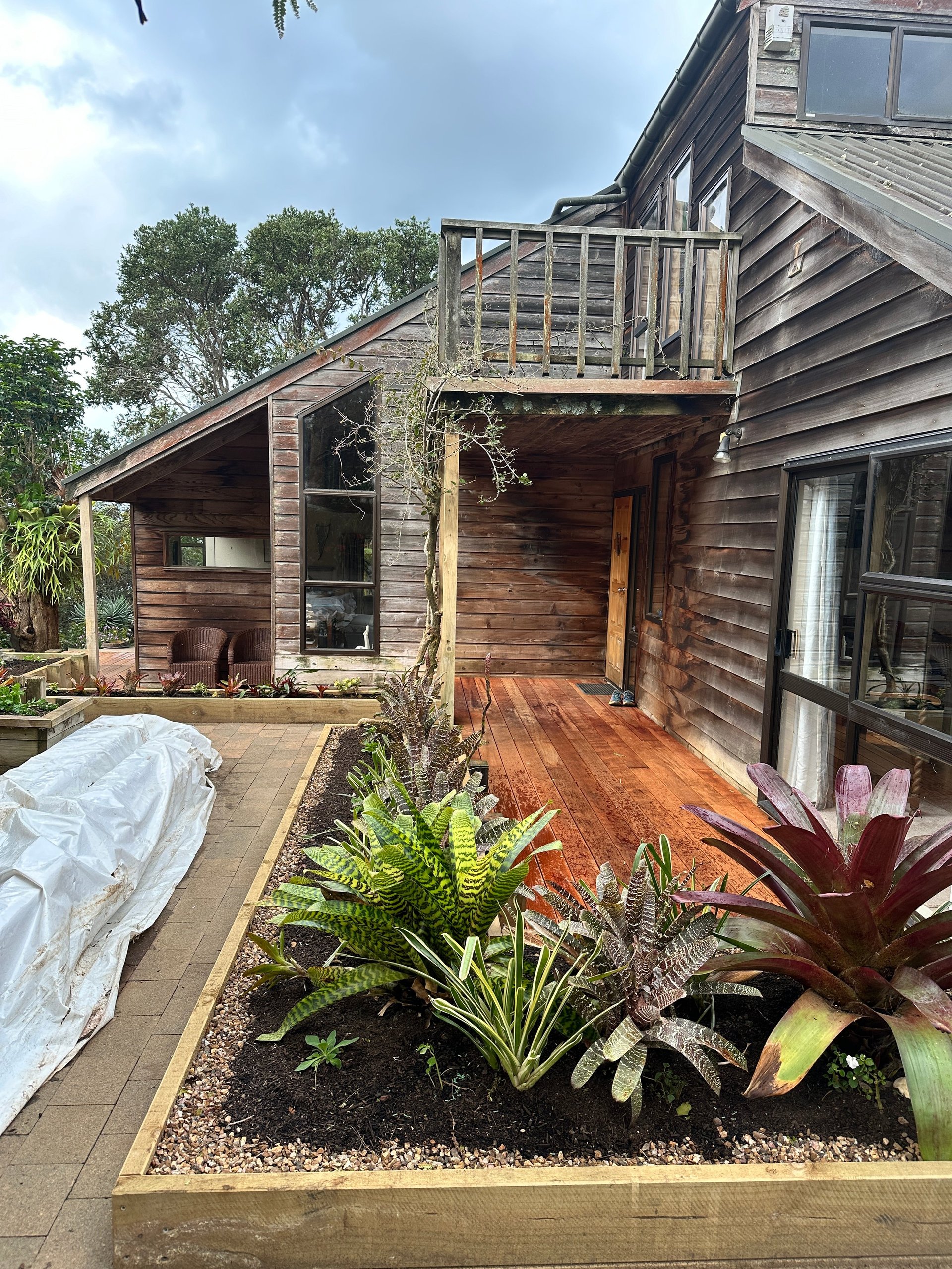 Modern wooden house with deck and upper balcony, surrounded by tropical plants and landscaping in front yard