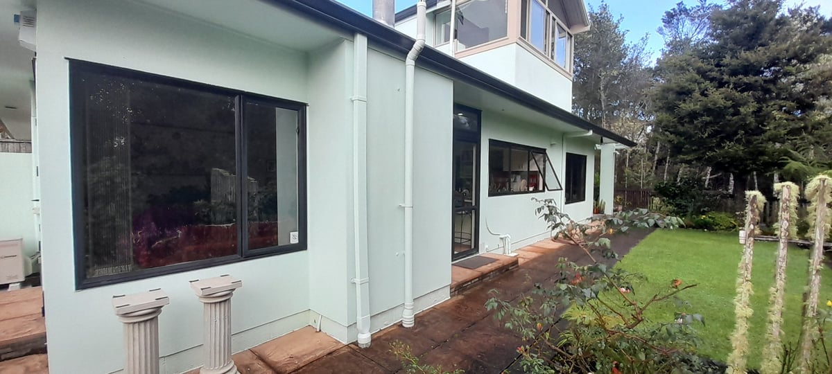 Modern light-colored house with large windows and wooden deck, surrounded by green lawn and wooden fence