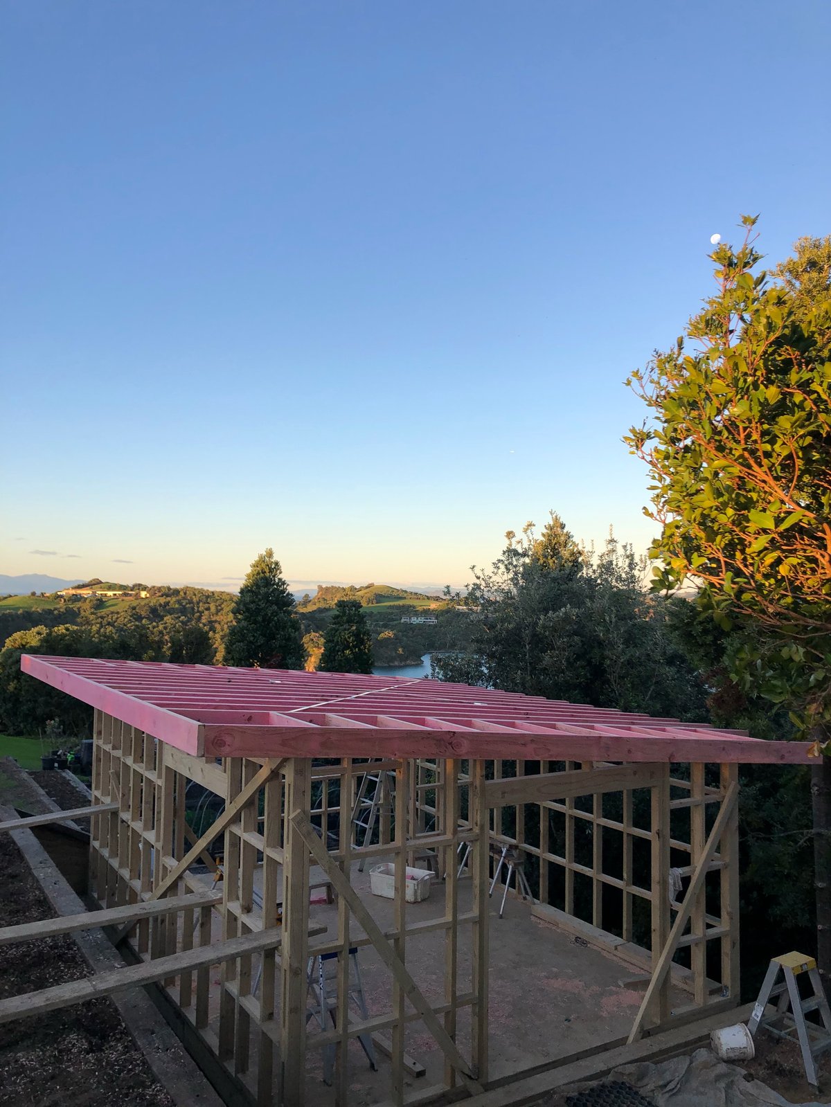 Timber frame structure under construction with burgundy metal roof, surrounded by hills and trees at golden hour