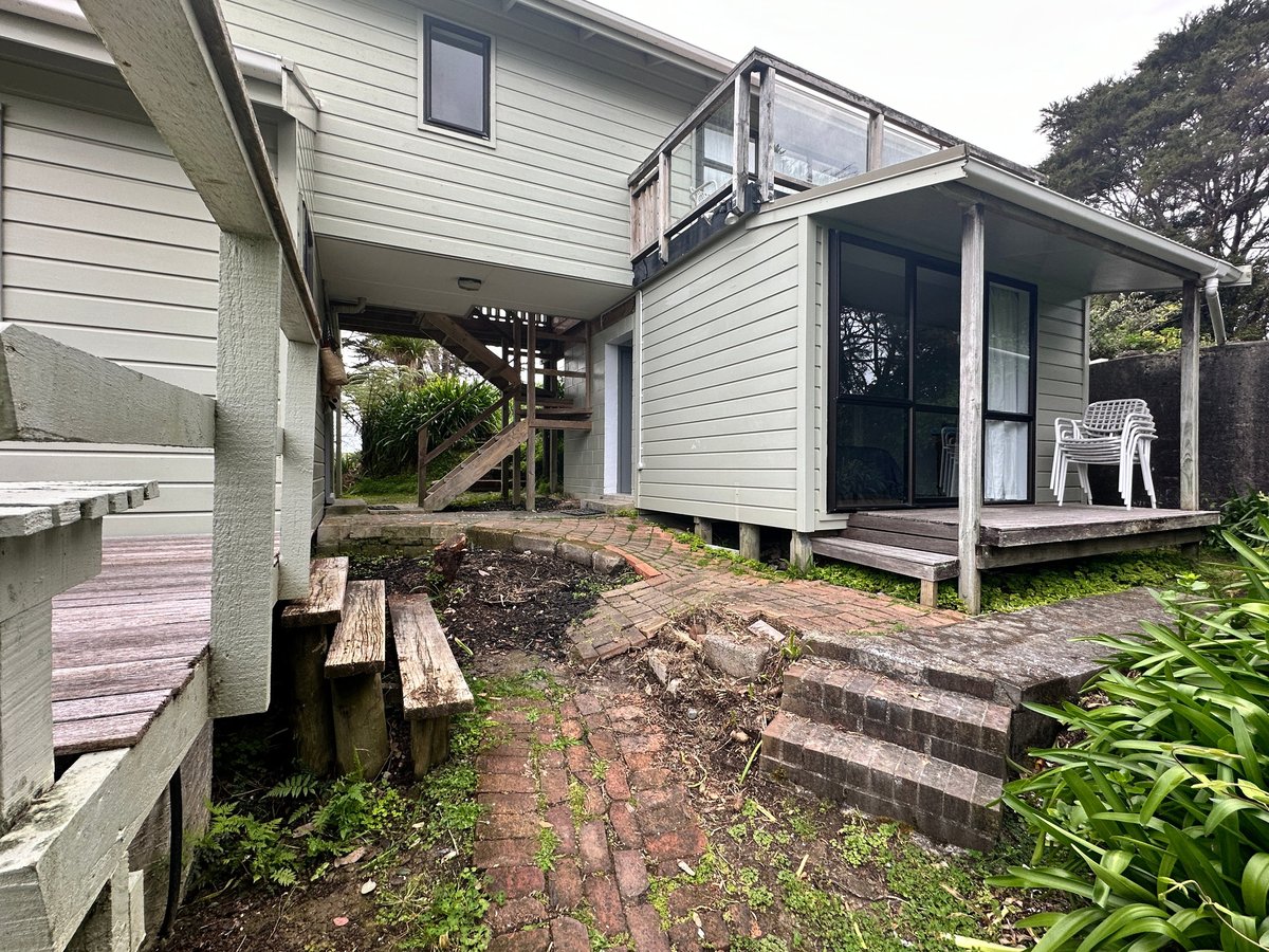 Elevated house on stilts with white siding, wooden stairs, and front deck with seating area surrounded by dirt and garden landscape