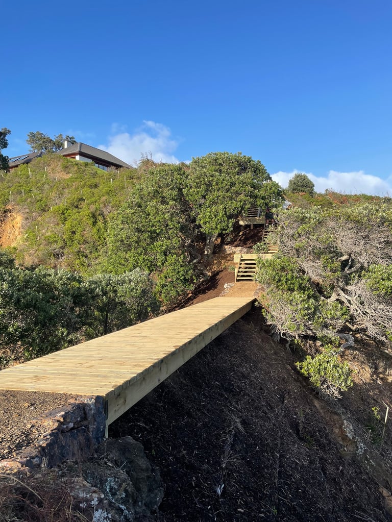 Wooden boardwalk section leading up a slope toward a clifftop house, with coastal vegetation on either side and a clear blue sky above