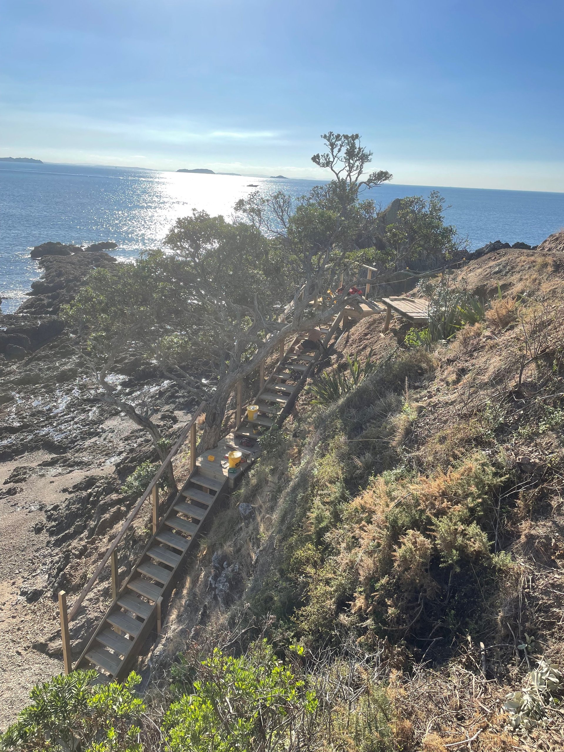 Long finished timber boardwalk with metal handrail descending a rocky coastal cliff toward the sea