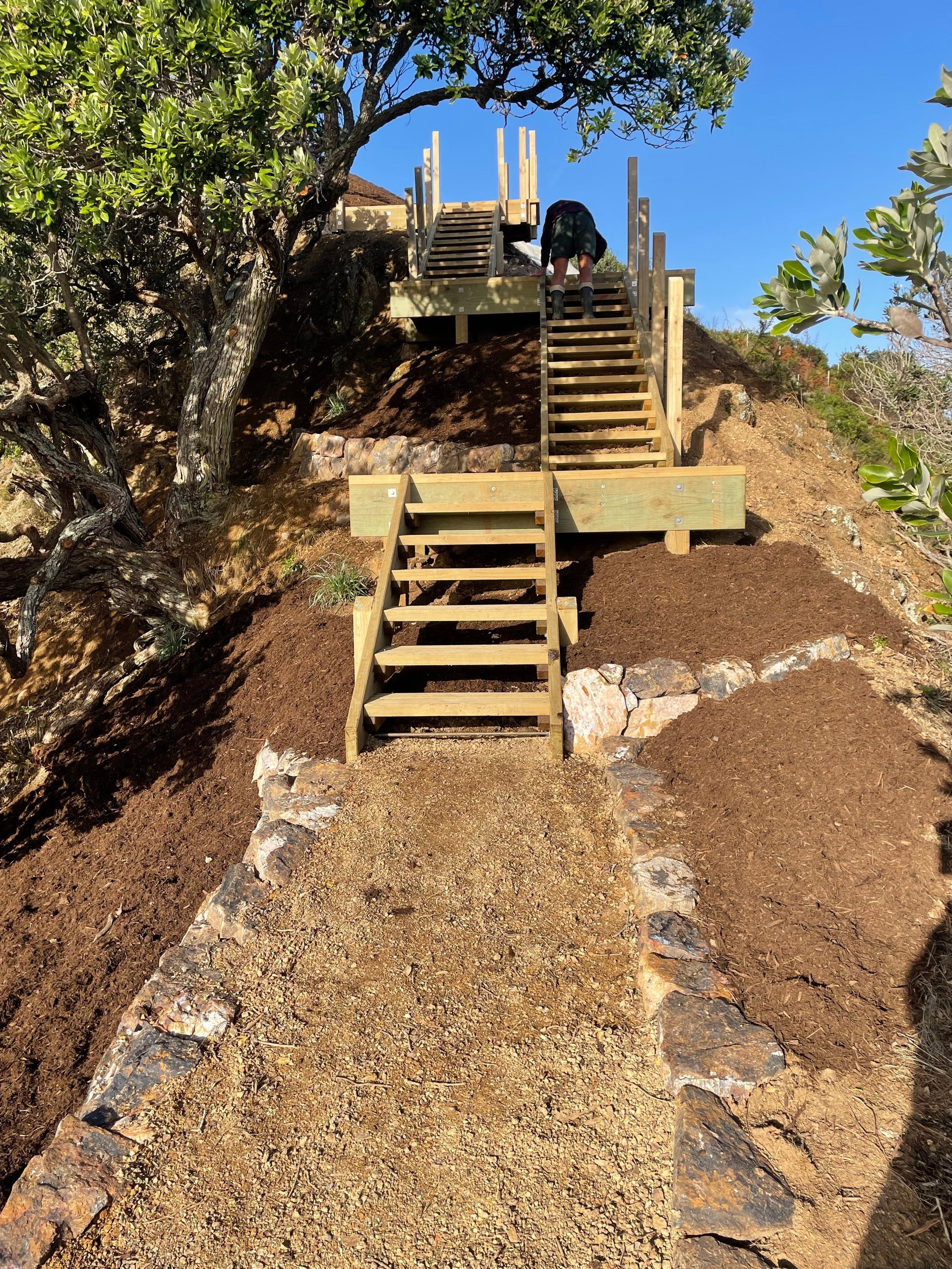 Wooden staircases and platforms built into a hillside path surrounded by trees and rocky terrain