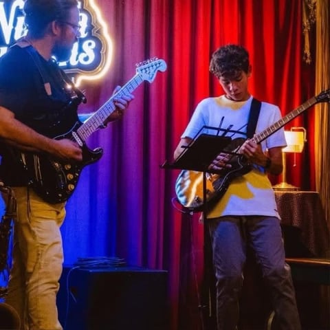 Two musicians performing with electric guitars on stage with red curtains and neon blue signage in background