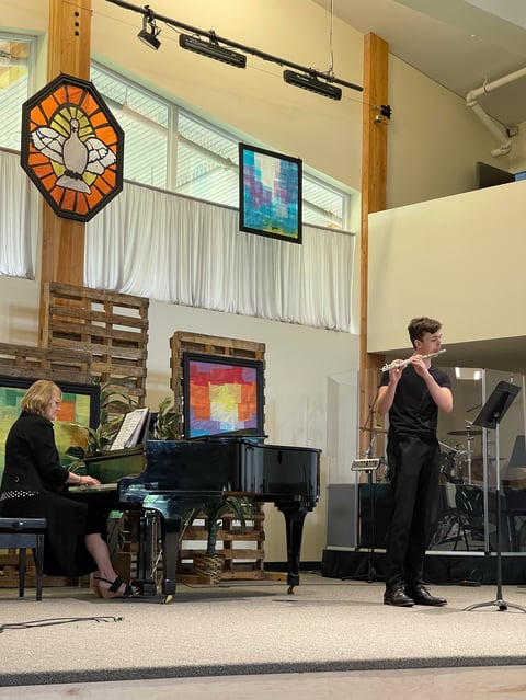 Two musicians performing on stage: a pianist in black on the left and a flutist in black on the right, with colorful stained glass artwork visible above