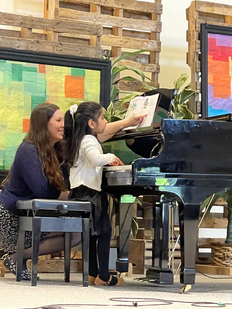 Two students sit at a piano reading sheet music during a lesson, with colorful panels and plants visible in the background