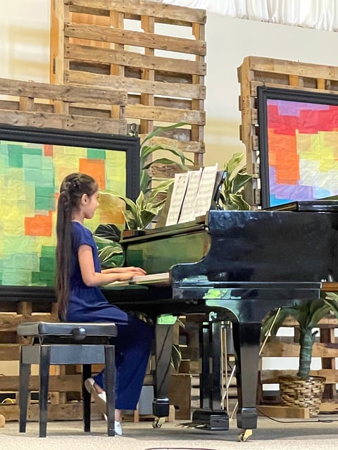 Young girl with long dark hair playing a black grand piano in front of colorful digital displays and wooden pallet backdrop