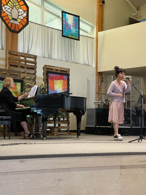 Woman singing at microphone on stage while pianist accompanies her in a church or concert hall with colorful window designs