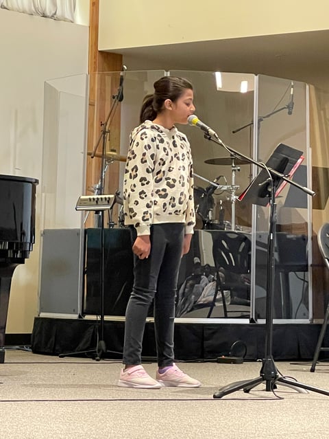 Young girl performing at microphone on stage wearing leopard print sweater and pink sneakers