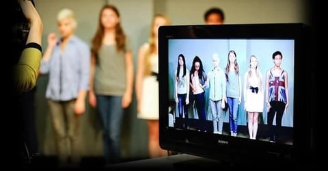 Video monitor displaying six people standing in a line while a group of people stand in the background out of focus