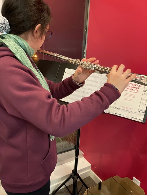 A musician in a maroon hoodie and glasses plays a flute while reading sheet music on a music stand against a red wall