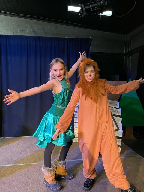 Two performers pose on stage with arms outstretched. One wears a turquoise dress and the other wears an orange costume with red hair and makeup