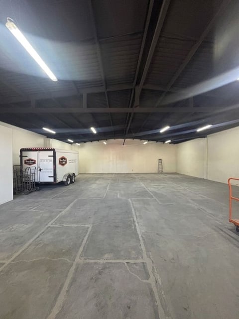 Almacén industrial vacío con piso de concreto, vigas expuestas, iluminación fluorescente y un pequeño remolque al fondo