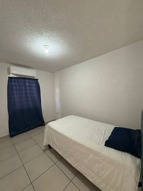 Recámara con cama individual, cortina azul oscuro, minisplit y piso de loseta