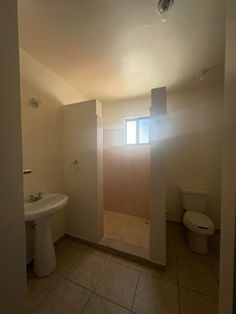 Baño vacío con lavamanos tipo pedestal, inodoro y regadera con cancel de vidrio bajo luz natural