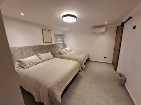 Recámara moderna con cama queen, ropa de cama blanca, cabecera beige, foco de techo y minisplit