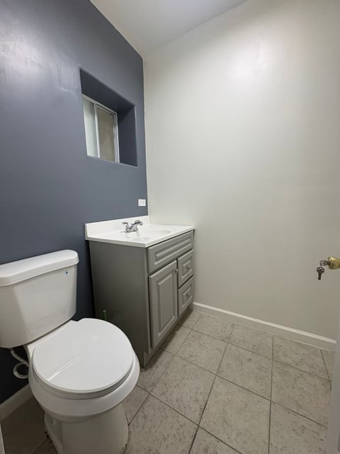Baño moderno con inodoro blanco, vanity gris con lavabo, pared acento azul marino, espejo y piso cerámico