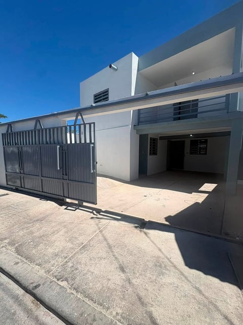 Casa blanca con portón metálico gris abierto mostrando cochera y balcón bajo cielo azul