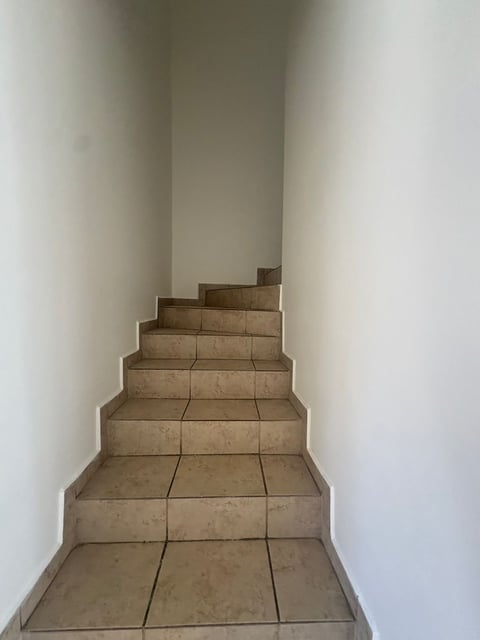 Escalera angosta con escalones de loseta beige ascendiendo entre paredes blancas