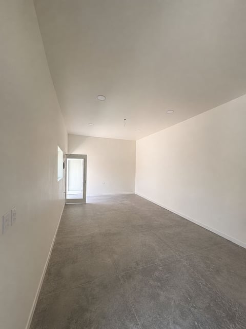 Sala vacía con paredes blancas, piso de concreto, luces empotradas en techo y entrada con puerta de vidrio al fondo