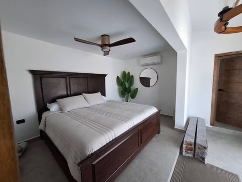 Recámara principal con cama king size de madera oscura, ropa de cama blanca, abanico de techo, planta y minisplit
