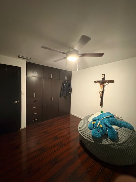 Recámara con clóset oscuro, ventilador de techo, piso de madera y crucifijo en la pared