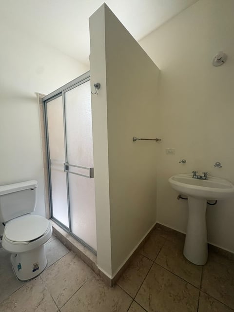 Baño completo con inodoro a la izquierda, regadera con cancel de vidrio al centro y lavamanos tipo pedestal a la derecha, piso de loseta beige