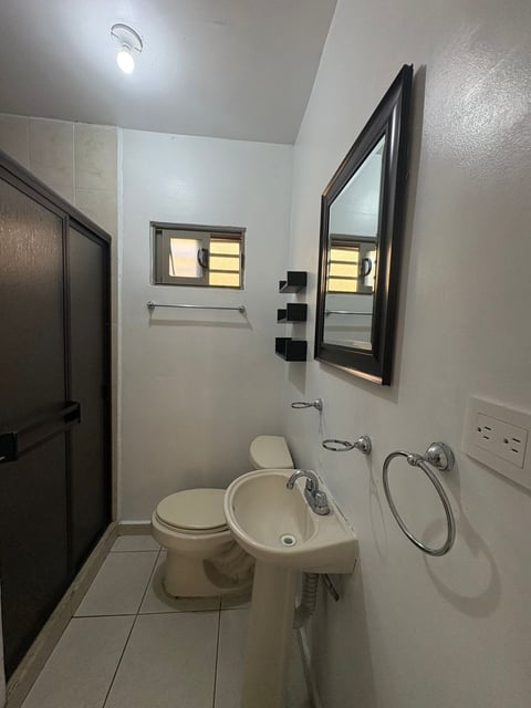 Baño moderno con paredes blancas, lavabo tipo pedestal, inodoro, espejo con marco negro, ventana pequeña y piso de loseta