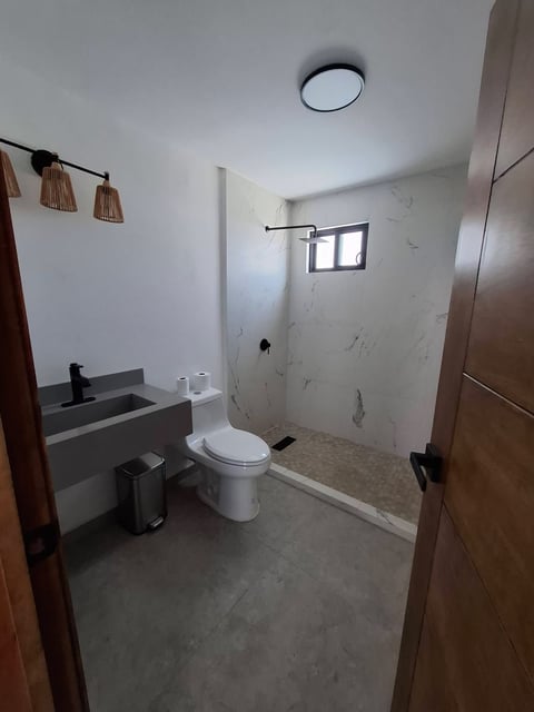Baño moderno con acabados en blanco y concreto, inodoro, lavabo, área de regadera con ventana, focos colgantes y puertas de madera