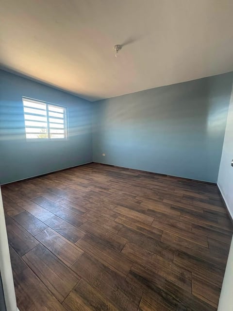 Recámara vacía con paredes gris azulado, piso de madera, ventana con persianas y foco en techo