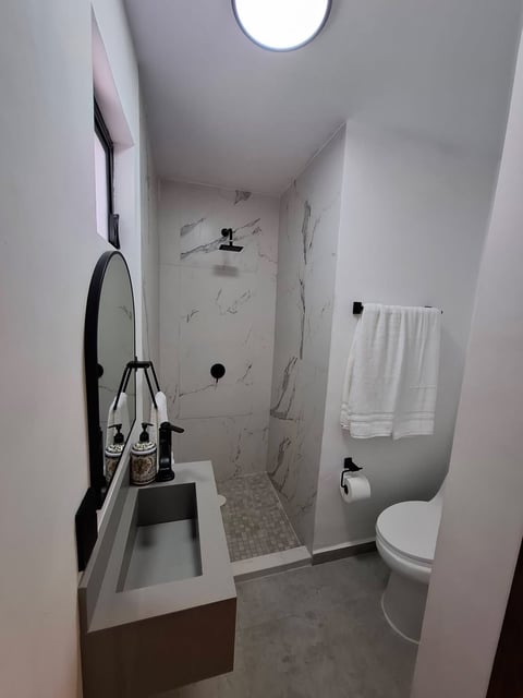 Baño moderno con regadera tipo walk-in, vanity blanco con lavabo, inodoro y foco circular en techo