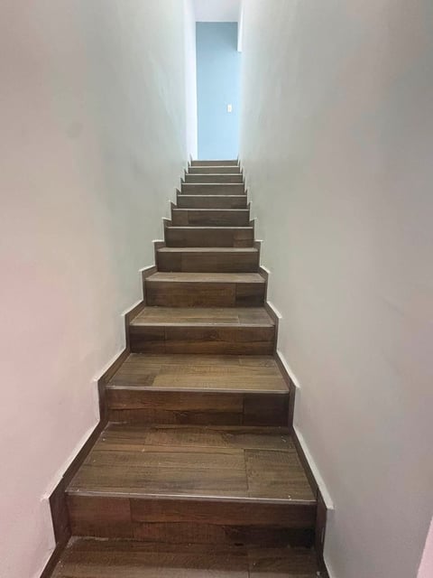Escalera larga con escalones de madera ascendiendo hacia luz azul al final, flanqueada por paredes blancas